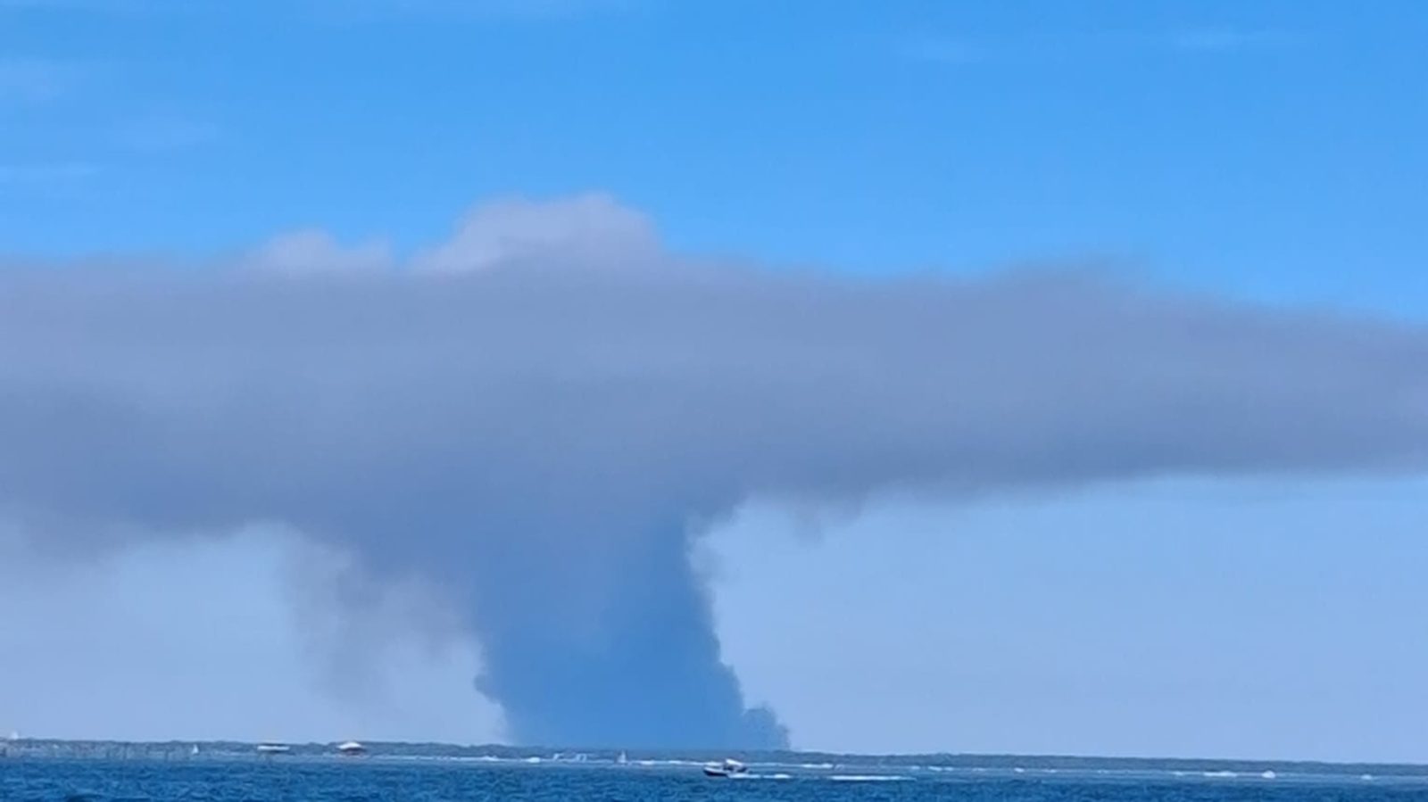 Un épais panache de fumée s’élève au-dessus du littoral et se couche en nappe sous un ciel bleu, vu depuis la mer.